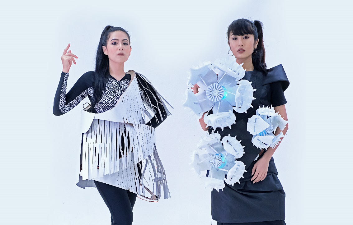 Two models posing with large wearable objects attached to their clothes